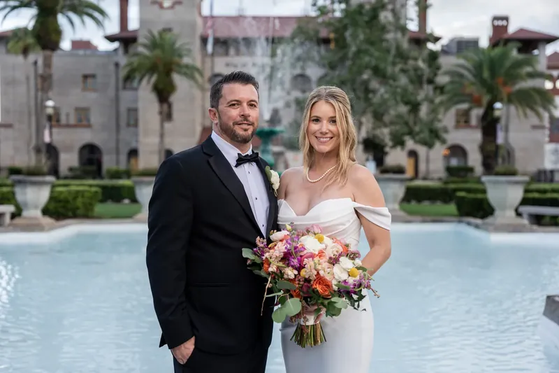 Jenna and Russell stand by a fountain in Downtown St Augustine, she holding a colorful bouquet in a white off-shoulder gown.