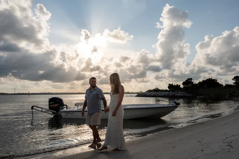 Jenna in a white dress and Russell in a blue shirt walk alongside their beached boat with dramatic clouds overhead.