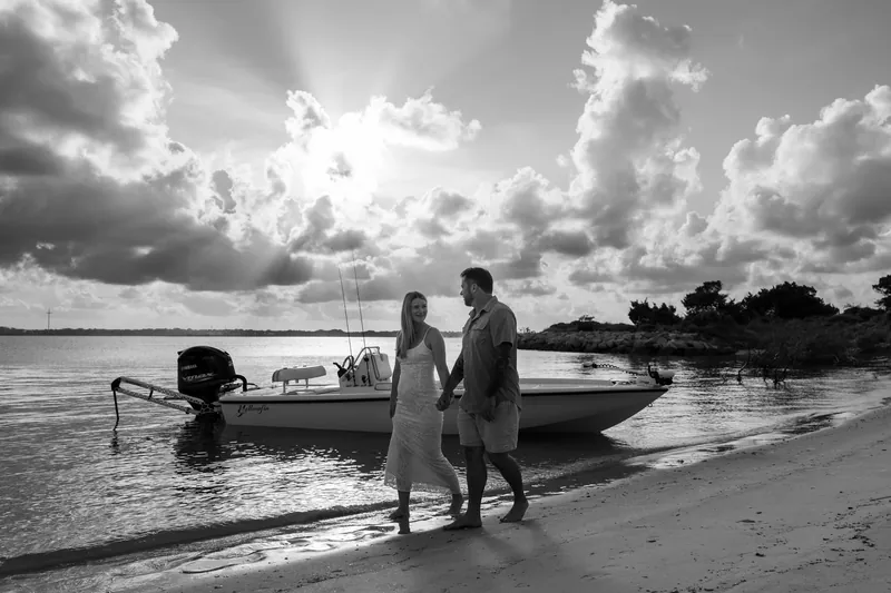 Black and white portrait of Jenna and Russell standing in shallow water next to their moored boat beneath towering clouds.