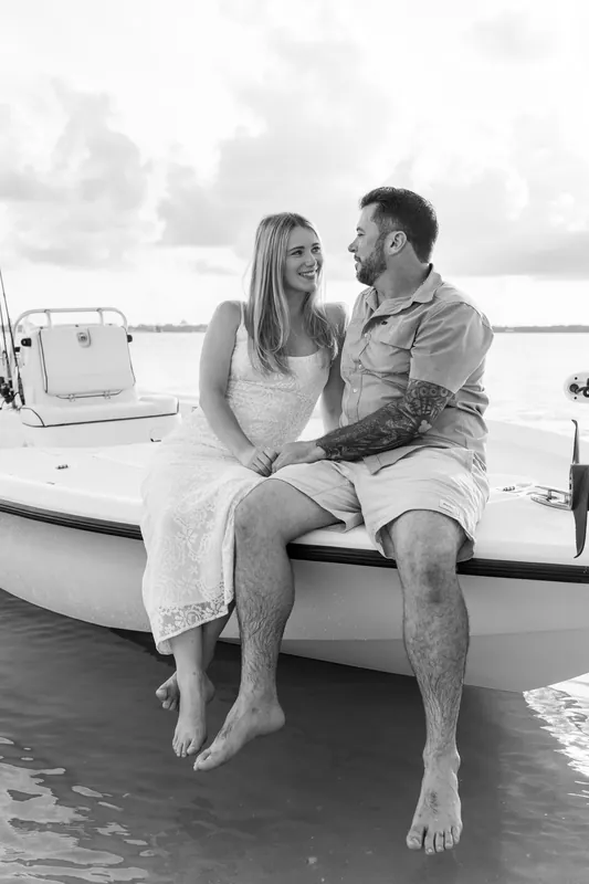 Black and white shot of the couple sitting barefoot on the boat, smiling at each other in profile.