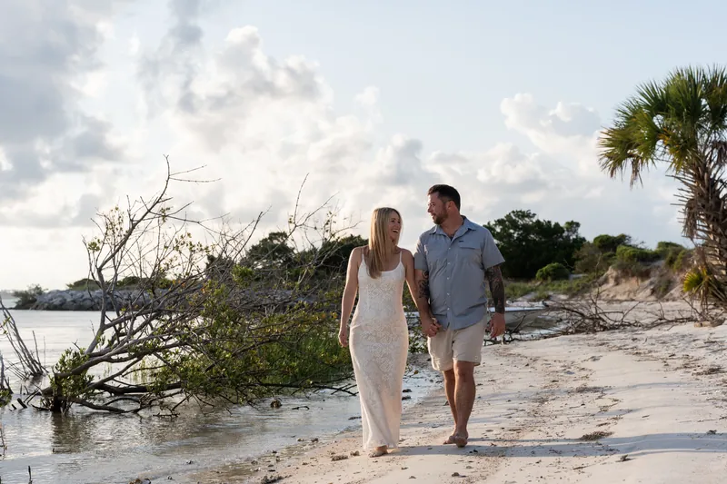 The pair walks hand in hand along the sandy shore at Matanzas Inlet, with driftwood and palmetto plants framing the inlet.