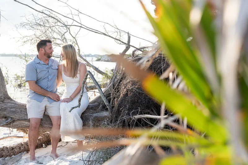 Russell and Jenna stand among weathered branches near the water's edge, with green foliage in soft focus.