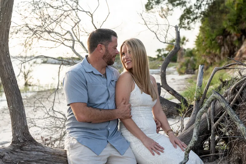 Russell and Jenna embrace on a sandy shore lined with gnarled trees at Matanzas Inlet, their figures framed against the water