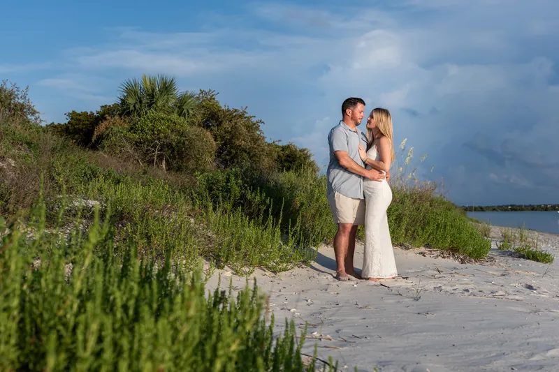 Jenna in a white dress and Russell in khaki stand close together on the beach at Matanzas Inlet under a blue sky dotted