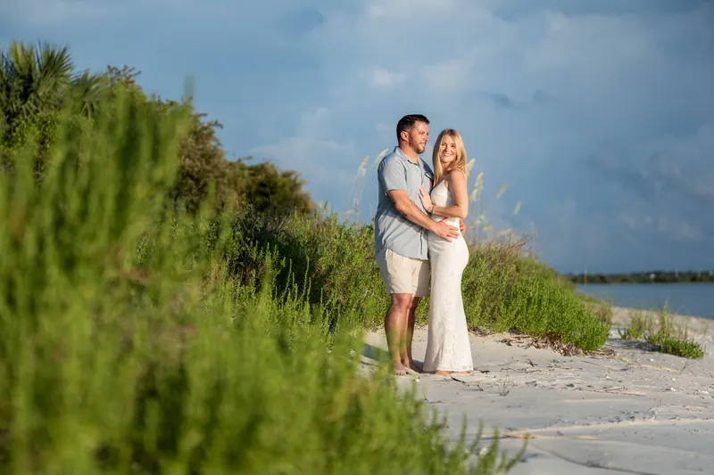 The couple stands on sand with marsh grasses in the foreground, the waterway and sky visible behind them at Matanzas Inlet.