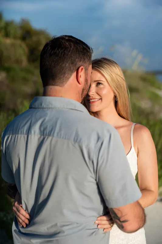 Russell holds Jenna close while she grins over his shoulder, the inlet's landscape soft and green behind them.