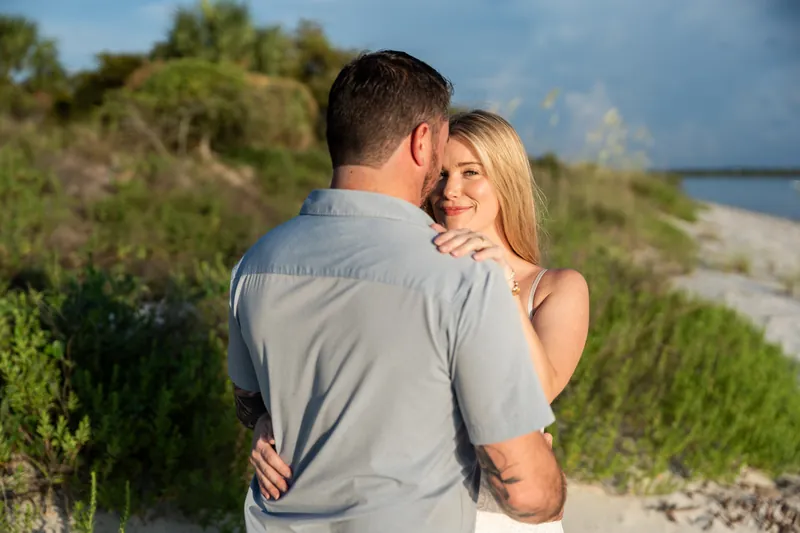 With his back to the camera, Russell hugs Jenna, who wraps her arms around him as coastal marsh stretches along the shoreline