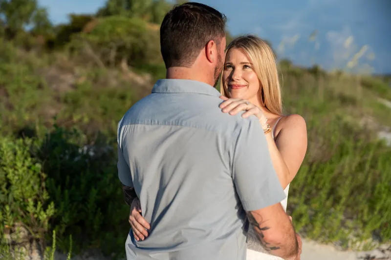 Russell embraces Jenna from behind as coastal grasses and blue sky frame them at Matanzas Inlet.