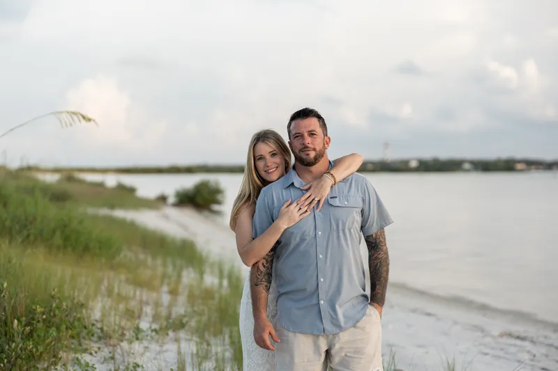 Russell and Jenna stand close on the beach with salt marsh and water stretching behind them.