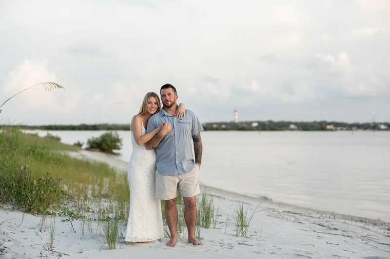 Jenna in a flowing white dress stands beside Russell in a gray shirt on the sand overlooking the inlet.