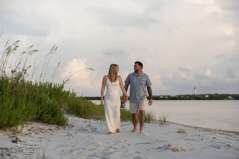 Jenna wears a white dress and Russell wears a blue shirt while walking along the sandy shore at Matanzas Inlet with tall