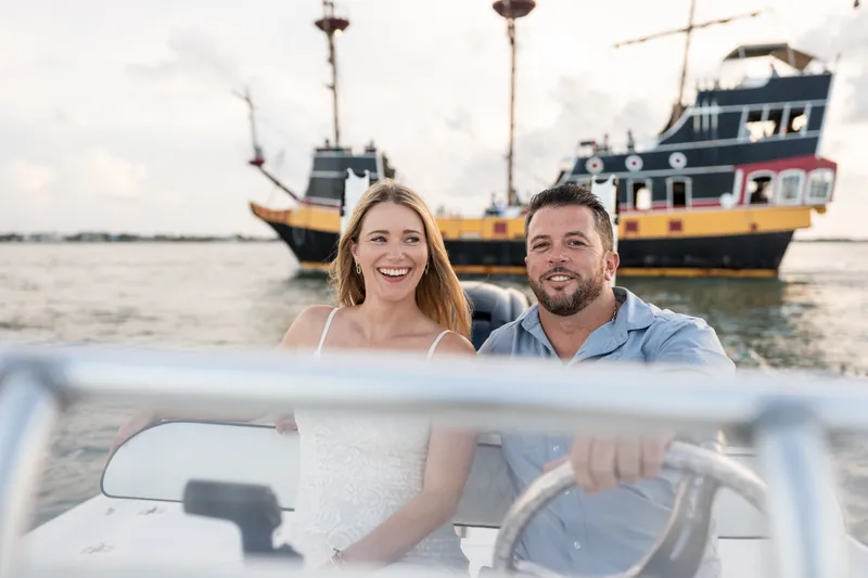 Jenna and Russell smile at each other from inside a boat at Matanzas Inlet, with fishing vessels anchored behind them.