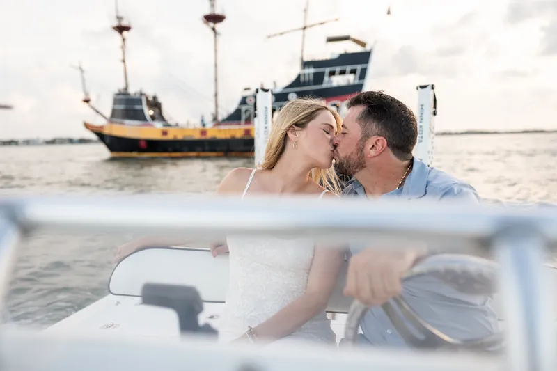 Russell kisses Jenna's cheek as they sit together on a boat, the inlet's moored ships visible across the water.