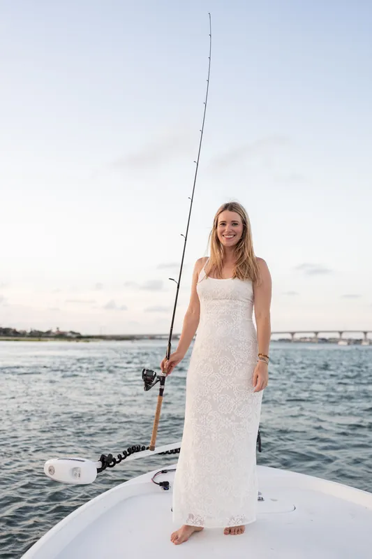 Jenna stands alone on the boat's bow in a white dress, fishing rod ready, the inlet stretching behind her.