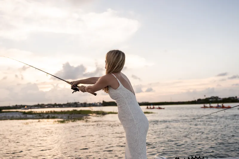 Holding a fishing rod, Jenna casts into the water at sunset, her white gown flowing as she stands on the boat.