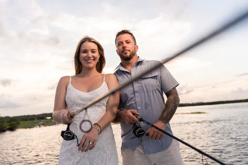 Russell and Jenna grip fishing rods together on the boat deck, sharing a relaxed moment under an overcast sky at the inlet.