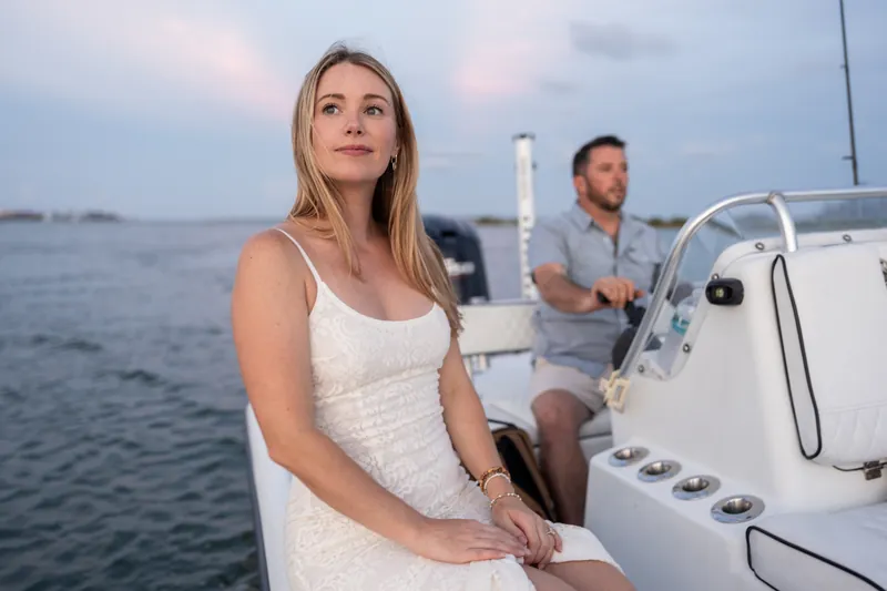 Jenna sits at the helm of a sailboat at dusk while Russell stands behind her, the calm water and distant shoreline stretching