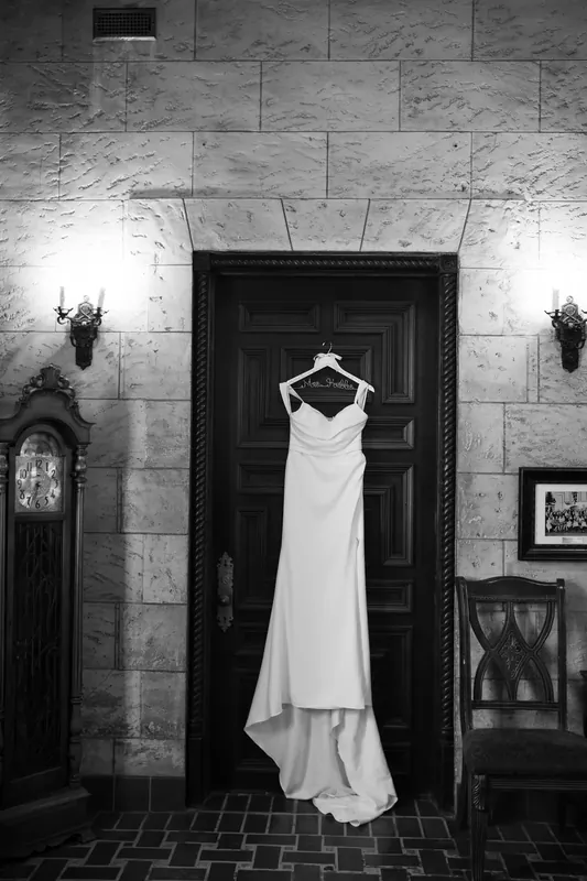 A white gown with layered skirt hangs against a dark wooden door on a marble-walled foyer in black and white.