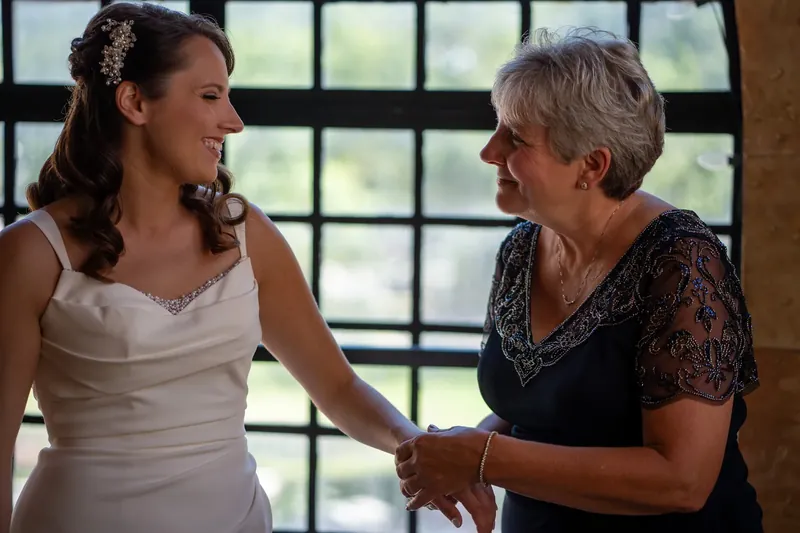 Jessica in her white wedding dress holds hands with an older woman in a black lace top, sharing a quiet moment by a window.