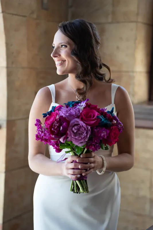 Jessica holds a vibrant bouquet of magenta and purple roses while standing in a stone archway at The Treasury on the Plaza.