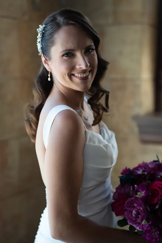 Jessica smiles at the camera wearing a white sleeveless gown with a floral hair clip and holding a deep purple bouquet.