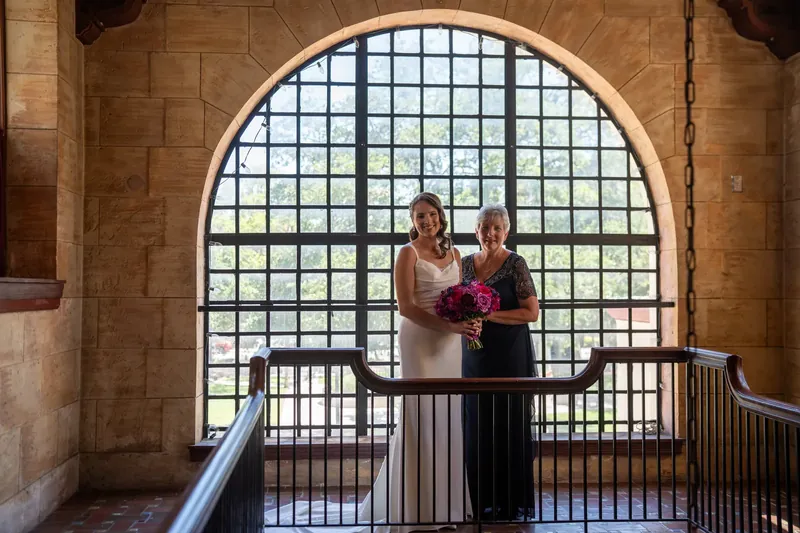 Jessica and John stand on a balcony beneath an arched window at The Treasury on the Plaza, holding flowers.