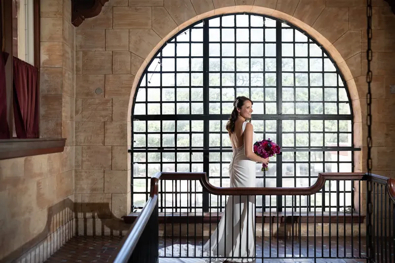 Jessica in her white wedding dress holds her purple bouquet on the balcony's arched window backdrop.