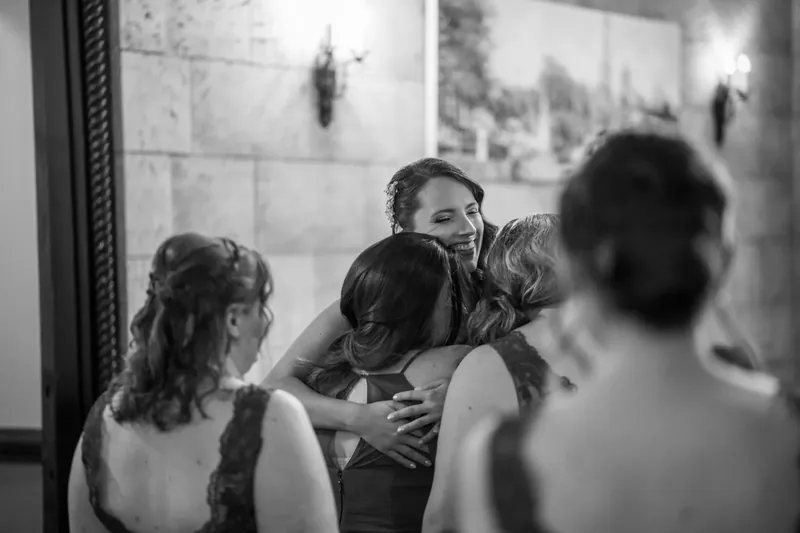 A man adjusts Jessica's necklace while she sits surrounded by bridesmaids in a candid black-and-white moment.