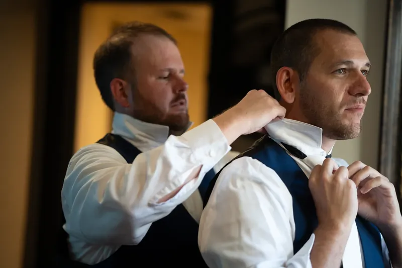 One man adjusts another's tie and vest in an indoor preparation moment.