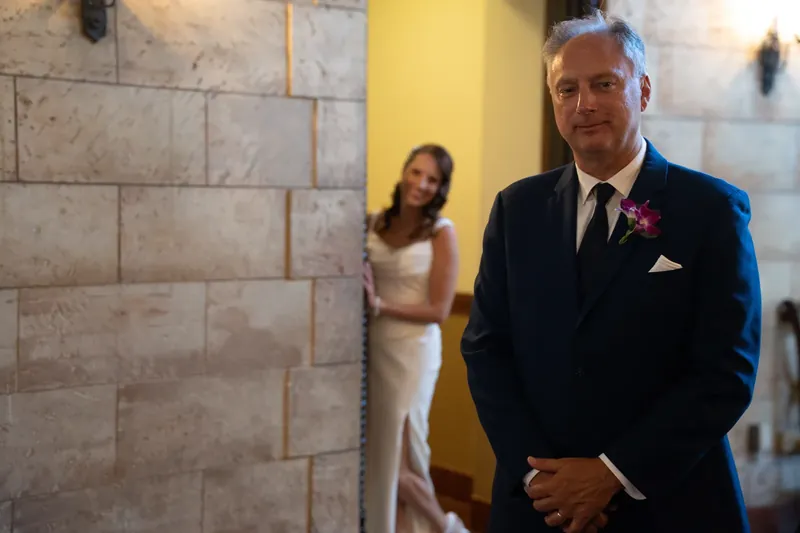 John stands in a navy suit near stone archway walls as Jessica waits in the background.