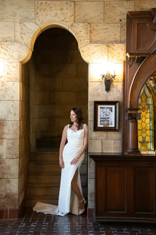 Jessica stands in her white wedding gown against the stone arches and wooden door of The Treasury on the Plaza.