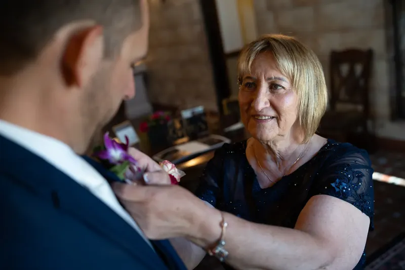 A woman in a dark sequined dress adjusts John's boutonnière as he looks on.