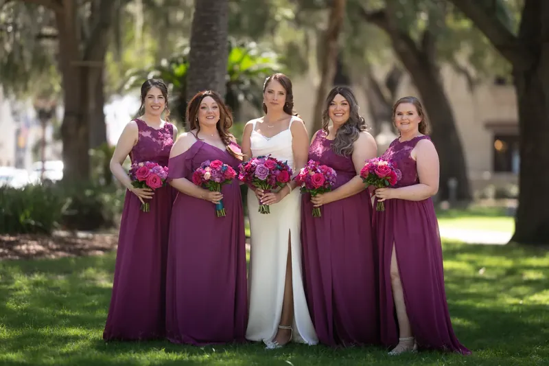Jessica stands center in white with four attendants in plum dresses holding deep pink bouquets beneath live oaks.