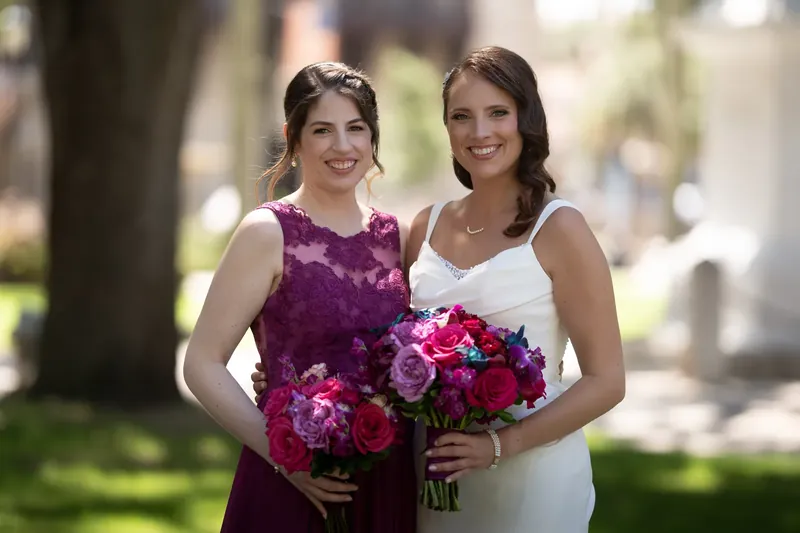 Jessica and an attendant pose together under dappled shade, both holding coordinating magenta and blush bouquets.