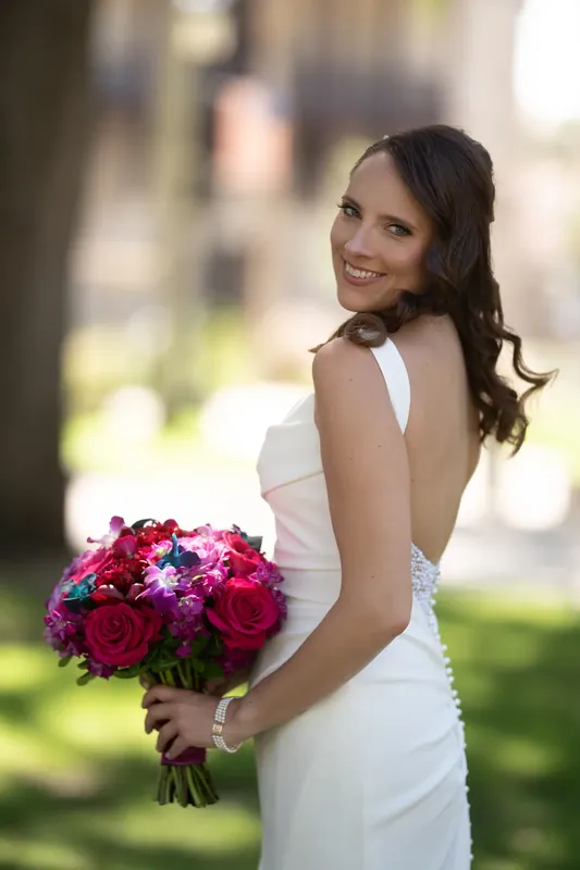 Jessica glances over her shoulder in her white gown, holding a fuchsia and burgundy bouquet in dappled sunlight.