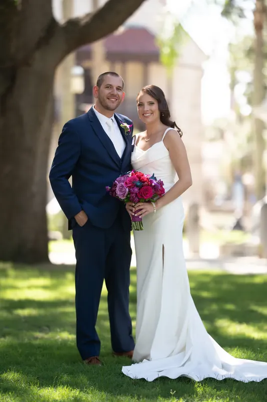 John in a navy suit and Jessica in white stand close together holding her pink bouquet, tree branches framing them.