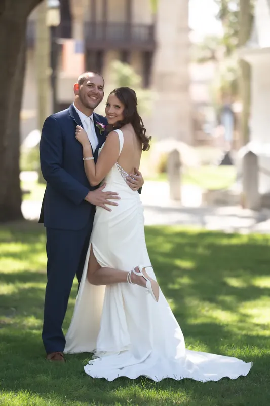 John embraces Jessica from behind as she holds her bouquet, both smiling under the shade of mature oaks.