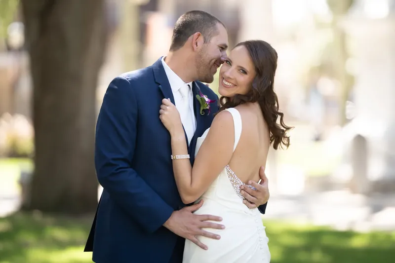 John kisses Jessica's temple while she smiles, his hand on her back as they stand together under tree cover.