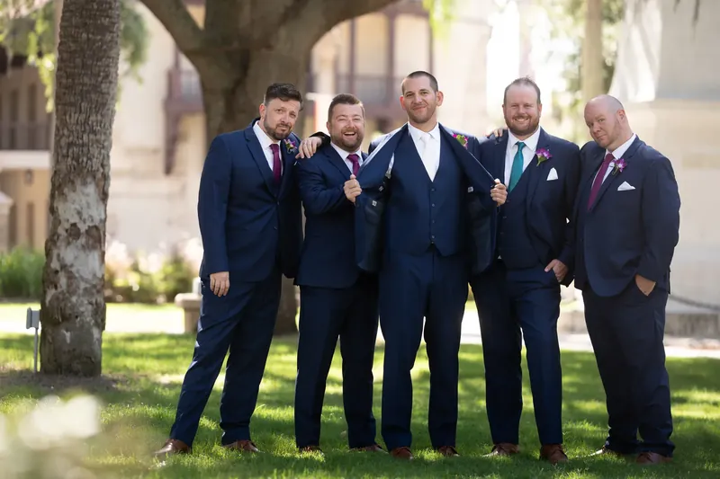 Five men in navy suits with pocket squares stand together under a tree at Plaza de la Constitución, smiling at the camera.