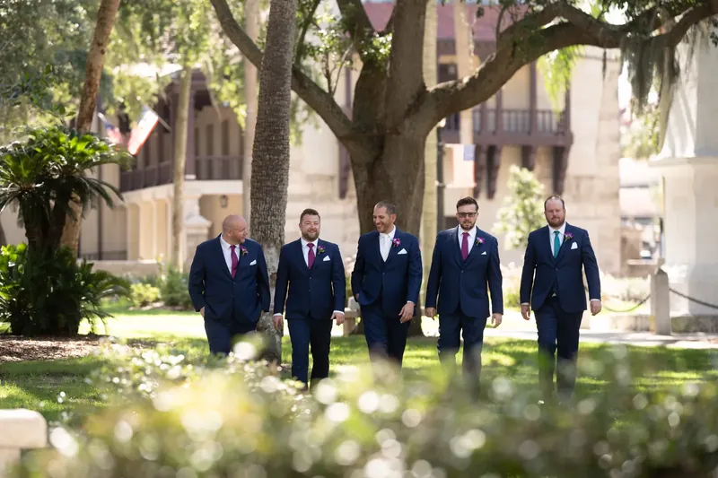John walks with four men in matching navy suits across a sunny plaza lawn bordered by historic buildings.