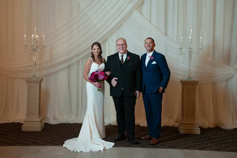Jessica in a flowing white gown holds a magenta bouquet while standing between two men in dark suits beneath draped fabric.