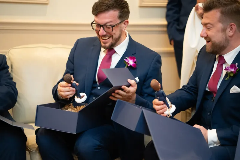 Two men in navy suits with pink ties laugh while opening small gift boxes during a groomsmen moment indoors.
