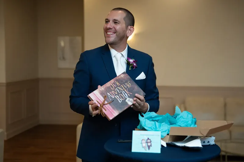John smiles while holding a wrapped gift and looking at items on a table decorated with turquoise fabric.