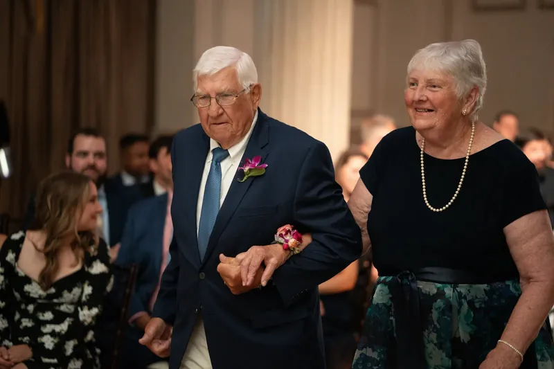 An older man in a navy blazer and a woman in a black dress walk arm-in-arm down the aisle.