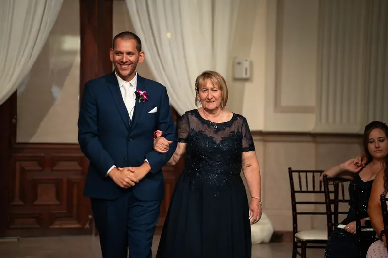 John, wearing a dark suit with a boutonnière, escorts a woman in a black beaded dress through the venue.