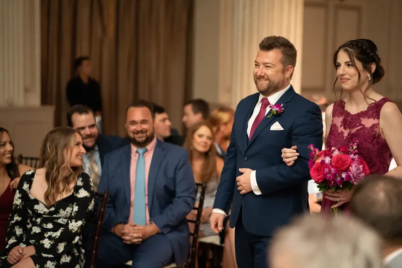 John walks down the aisle holding a bouquet, smiling beside a woman in a burgundy dress as guests watch.