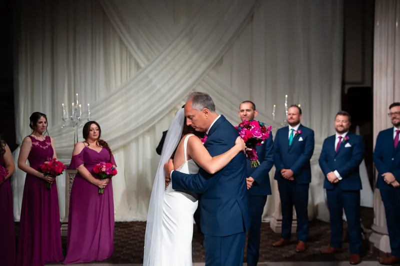 John dips Jessica in an embrace while bridesmaids in magenta and groomsmen in navy suits look on during their first dance.