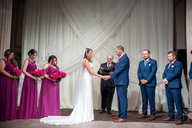 Jessica in white stands beside John in navy while an officiant reads their vows, with attendants flanking them.