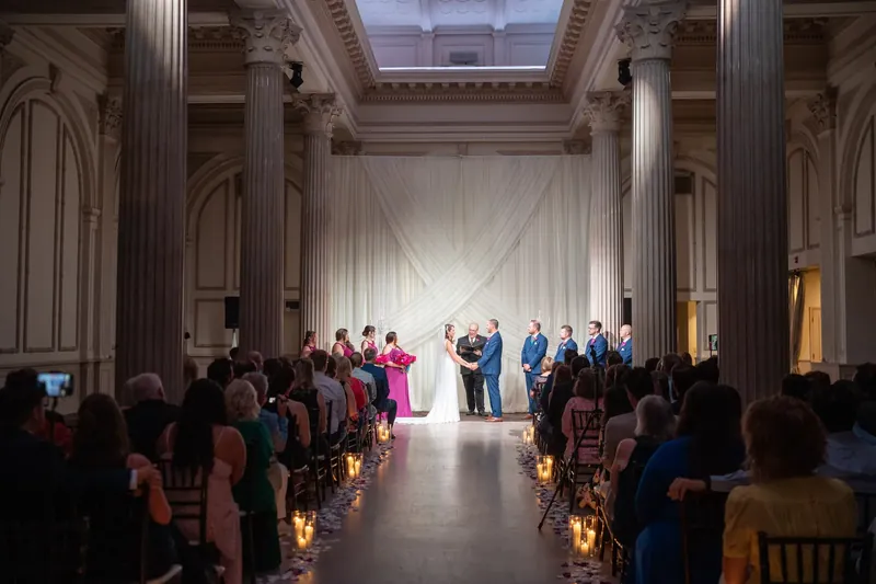 The couple walks down the aisle together through the columned ballroom lined with candlelit aisles and seated guests.