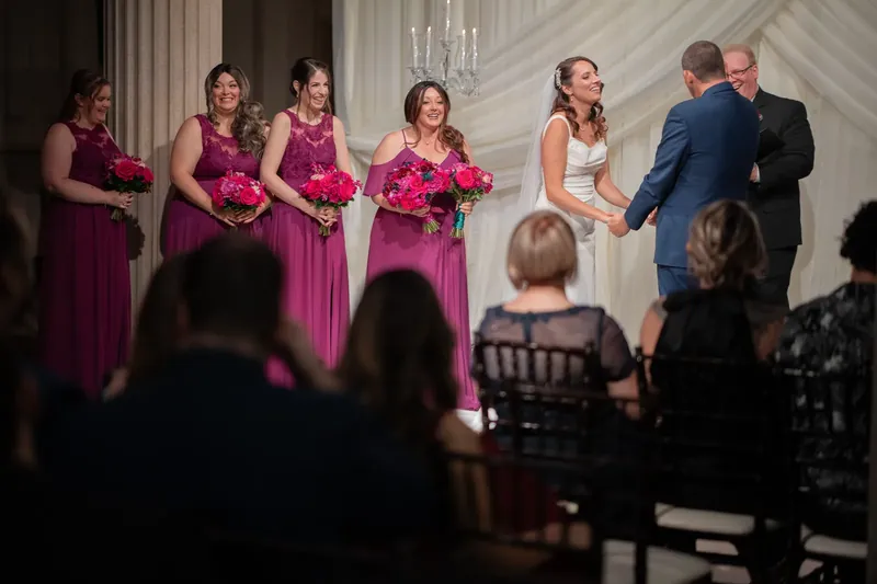 Jessica stands with five bridesmaids in matching magenta dresses at The Treasury on the Plaza, flanked by formal columns.